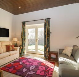 A living room with two sofas a wall mounted television a floral area rug and glass doors with curtains at Stable Cottage in Triscombe
