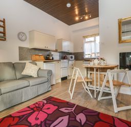 A living area with a grey sofa green cushion a small dining table with two chairs kitchen cabinets appliances and a wall-mounted electric fireplace at Stable Cottage in Triscombe