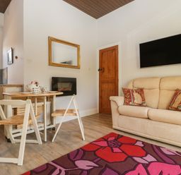 A living room with a beige sofa and two red pillows a wall mounted TV a wooden door a small wooden table with chairs a floral rug a mirror and a kitchen area at Stable Cottage in Triscombe