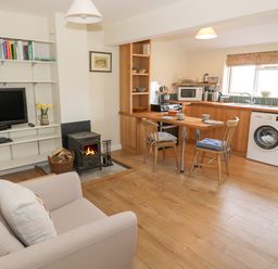 An open plan living room and kitchen with a sofa wood stove dining table and chairs and shelves with books at The Wash House in Sibford Ferris