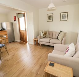 A living area with two sofas, a wooden door, framed pictures on the wall, and a small kitchen with appliances at The Wash House in Sibford Ferris