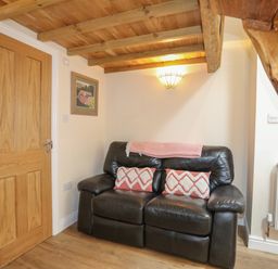 A small living area with a black leather sofa two patterned cushions a pink throw blanket a wooden door wooden ceiling beams and stairs at Glan Meon Cottage in Barmouth