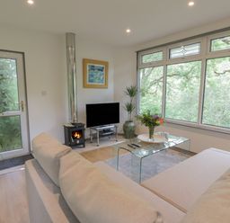 A living room with a beige sofa glass coffee table plants a fireplace and large windows at The River Lodge in Oban