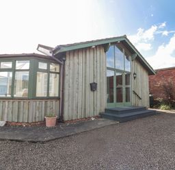 A wooden building with large windows and green frames next to a gravel area at The Shieling in Warkworth