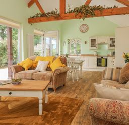 A sitting area with upholstered sofas yellow cushions wooden coffee table and a kitchen in the background at Langdale Barn in Sampford Courtenay