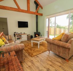 A living room with sofas a coffee table a wall-mounted TV a wood-burning stove and large sliding glass doors at Langdale Barn in Sampford Courtenay