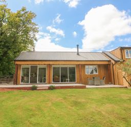 A wooden house with sliding glass doors a patio table and chairs and a lawn at Langdale Barn in Sampford Courtenay