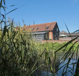 A house with a tiled roof near a pond with tall grasses in the foreground at Kingfisher 2 The Old Stables Witney