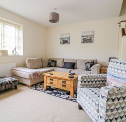 A living room with a beige sectional sofa, a wooden coffee table, a patterned armchair, and paintings of boats on the wall at 26 Burtons Mill in Stalham