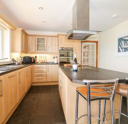 A kitchen with wooden cabinets a black countertop a sink microwave oven and bar stools at Trevose House in Crantock