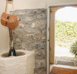 An interior stone wall with a large champagne bottle in a built-in niche next to an open door showing outdoor plants at Higher Roskorwell Cottage in St Keverne