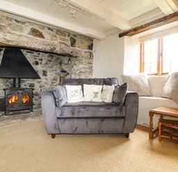 A living room with a stone fireplace and wood burning stove a gray sofa with cushions a window seat with pillows and a wooden side table at Higher Roskorwell Cottage in St Keverne