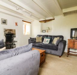 A living room with two gray sofas cushions a wooden chest an old stone fireplace a wooden TV stand and a television at Higher Roskorwell Cottage in St Keverne