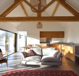 An open plan living room and kitchen with wooden beams a grey sofa a chair a dining table and large windows at High Cogges Farm Holiday Cottages - The Cart Shed in High Cogges near Witney