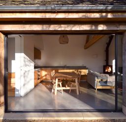 A kitchen and living area with a wooden dining table and chairs a gray sofa and a wood burning stove at High Cogges Farm Holiday Cottages - The Cart Shed in High Cogges near Witney