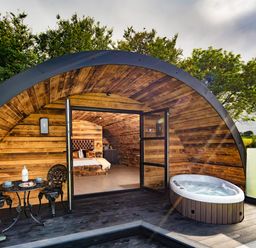 A wooden pod with an open door showing a bed inside a patio with a small table and chairs and a hot tub at The Piglet Pod in Llanarth