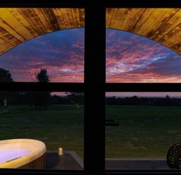A view through a window showing a lit hot tub on a deck and a grassy field with trees at sunset at The Piglet Pod in Llanarth