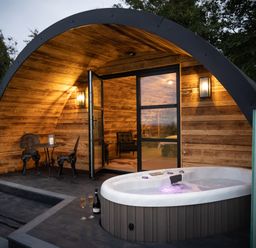 A wooden curved cabin with a hot tub two chairs and a small table at The Piglet Pod in Llanarth
