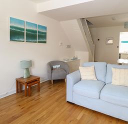 A living room with a light blue sofa two cream pillows a grey armchair a small wooden table with a lamp and a triptych wave painting on the wall at Gulls Nest in Pakefield