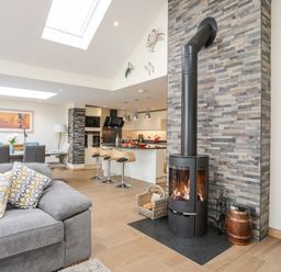 An open living room and kitchen area with a gray sofa two patterned pillows a wood burning stove with a stone chimney and a kitchen island with bar stools at The Herun in Rhosneigr