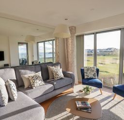 A living room with a gray sofa and blue chairs next to large windows overlooking a grassy area at Newquay Fistral Beach View in Newquay