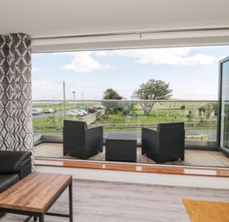 A lounge area with black chairs and a wooden table by large open glass doors leading to a balcony with a view of a grassy area at Amble Views in Amble