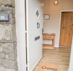 An open white door revealing a wooden floor inside a hallway with a wooden console table a hanging light and a door mat with Croeso at Y Stabl in Llanddoged near Llanrwst