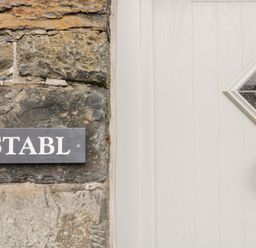 A stone wall with a wooden sign reading Y STABL and a cream door with a diamond glass window and black ring door knocker at Y Stabl in Llanddoged near Llanrwst