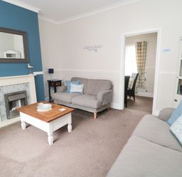 A living room with two sofas a wooden coffee table a fireplace a blue accent wall and a doorway leading to a dining area at Sandy Toes Cottage in Newbiggin-By-The-Sea