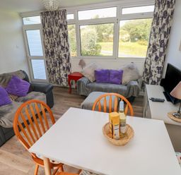A living room with two sofas purple cushions a white dining table wooden chairs a TV on a cabinet and windows with floral curtains at Y Twll Bollt (The Bolt Hole) in Folkeston Hill Nolton Haven West Wales & Pembrokeshire