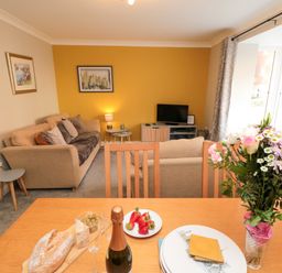 A living room with beige sofas a television and paintings on the walls with a dining table holding a bottle of champagne bread strawberries and flowers at Esk Retreat in Whitby