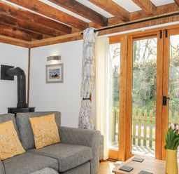 A living room with a gray sofa with yellow pillows a wood burner and glass doors opening to a garden at Stow Cottage in Davidstow near Camelford