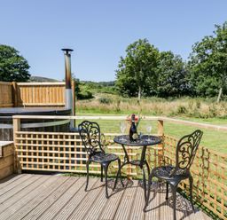 An outdoor wooden deck with a small black metal table and two chairs a hot tub with steps and wooden fencing with trees and grass in the background at Embden Pod at Banwy Glamping in Llanfair Caereinion