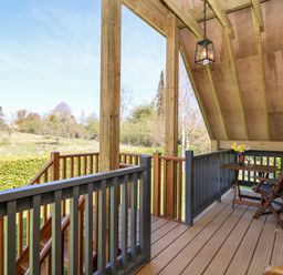 A wooden covered balcony with railing a small table and two chairs overlooking a grassy area and trees at Cherrywood Barn in South Petherton