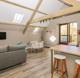 An open living space with a gray sofa a small dining table two white chairs kitchen counter with two stools and wooden beams on the ceiling at Cherrywood Barn in South Petherton