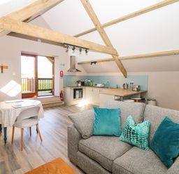 An open kitchen and living area with wooden beams a round dining table and a gray sofa with teal cushions at Cherrywood Barn South Petherton