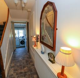 A hallway with a staircase and a shelf with a lamp clock mirror and vase leading to a kitchen area at Ty Coch y Castell in Pembroke