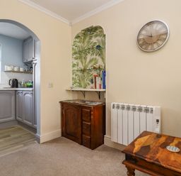 A room with a wooden cabinet under a recessed shelf with books and two glass shelves with plants and decor and a wall clock next to a kitchen with grey cabinets