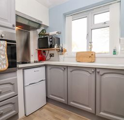 A kitchen with grey cabinets a small white fridge a microwave on a wooden shelf and a window above the counter at Molly's Bit in Church Westcote near Bourton-On-The-Water