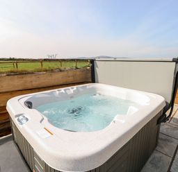 An outdoor hot tub with bubbling water on a stone patio next to a wooden fence with fields and hills in the background at Glowty Brynbedw in Efailwen