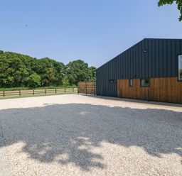 A gravel driveway next to a modern building with wood and metal exterior near trees and a wooden fence at Tilia Barn in Wincanton