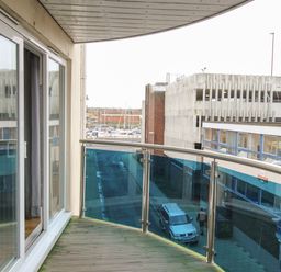 A balcony with glass and metal railing overlooking a street with buildings and parked cars at Nautical Breeze in Weymouth