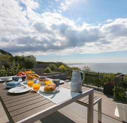 An outdoor dining table with breakfast items and drinks on a patio overlooking the sea at Sea Sands in Praa Sands