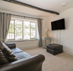 A living room with a leather sofa a window with curtains a side table with a lamp a wall-mounted TV and an ottoman at Pixie Cottage in Looe