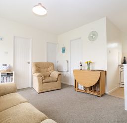 A living room with beige armchairs a wooden folding table with flowers and wine near a kitchen at 14 Siesta Mar in Mundesley