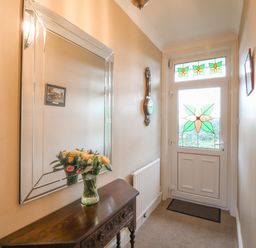 A hallway with a wooden table holding a vase of flowers a large mirror on the wall a clock a radiator and a door with stained glass at Silver Crooks in Trewyn Nr Holsworthy