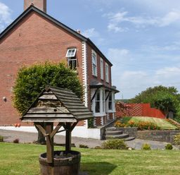 A brick house with white window frames and a wooden wishing well in the garden at Silver Crooks in Trewyn Nr Holsworthy