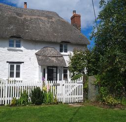 A white cottage with a thatched roof and a white picket fence surrounded by grass and greenery at Furneaux Hatch in Churchstow
