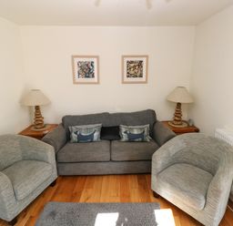 A sitting area with a gray sofa, two gray armchairs, two side tables with lamps, and framed pictures on the wall at Oysterbank Cottage in Llangwm Pembrokeshire