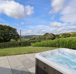 An outdoor hot tub on a stone patio overlooking a lawn and green hedges with hills and trees in the distance at Pen Bryn in Cyffylliog near Ruthin